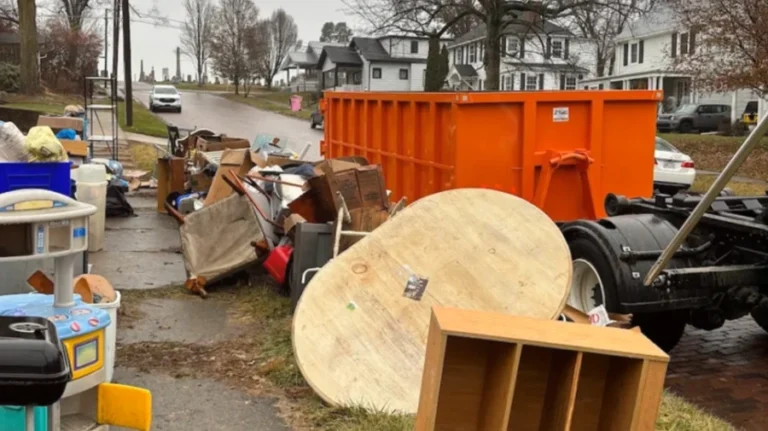 Curbside pile of old furniture and household junk beside an orange roll-off dumpster on a residential street