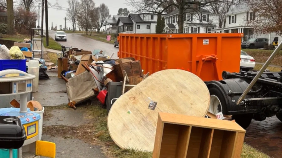 Curbside pile of old furniture and household junk beside an orange roll-off dumpster on a residential street